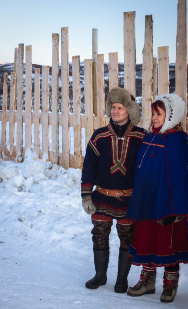 A male and female Sámi who greet you and are part of the whole experience. They are both dressed in their traditional Sámi clothing. There is snow covering the ground and a fence behind them.