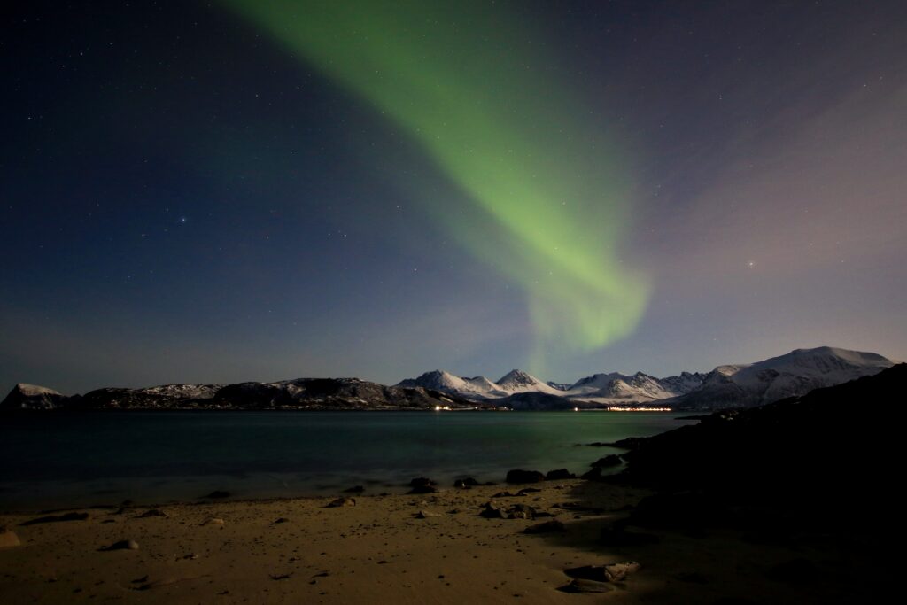 Photo Courtesy of Jiri Dvorak (NL Tour Guide). The beach and sea are in the foreground, with the snowy mountains and Northern Lights (Aurora) in the background.