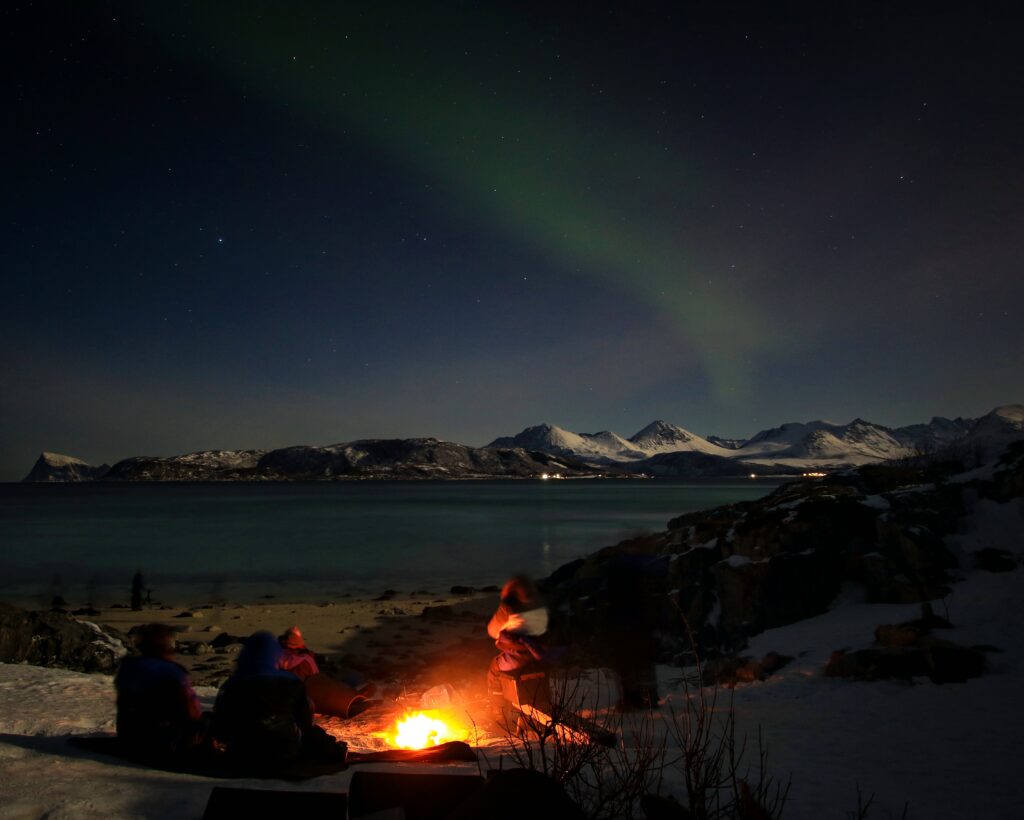 Photo Courtesy of Jiri Dvorak (NL Tour Guide). The beach, campfire (with some people sitting by the fire) and sea are in the foreground. The snowy mountains and Northern Lights (Aurora) in the background.
