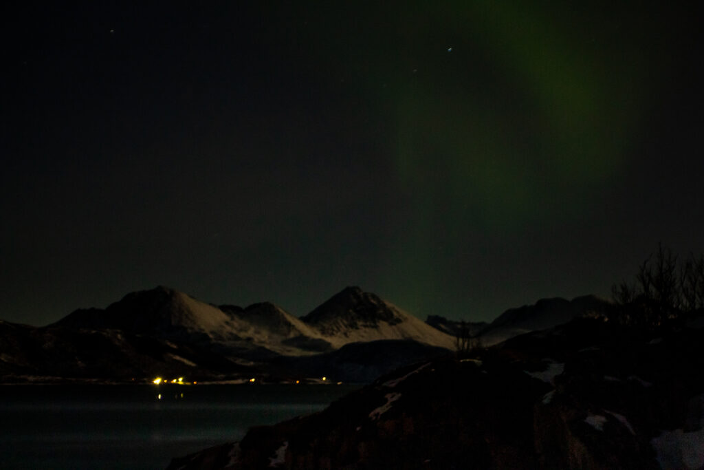 An image of the sea and some snowy mountain peaks below, with a hint of Aurora dancing across the dark sky.