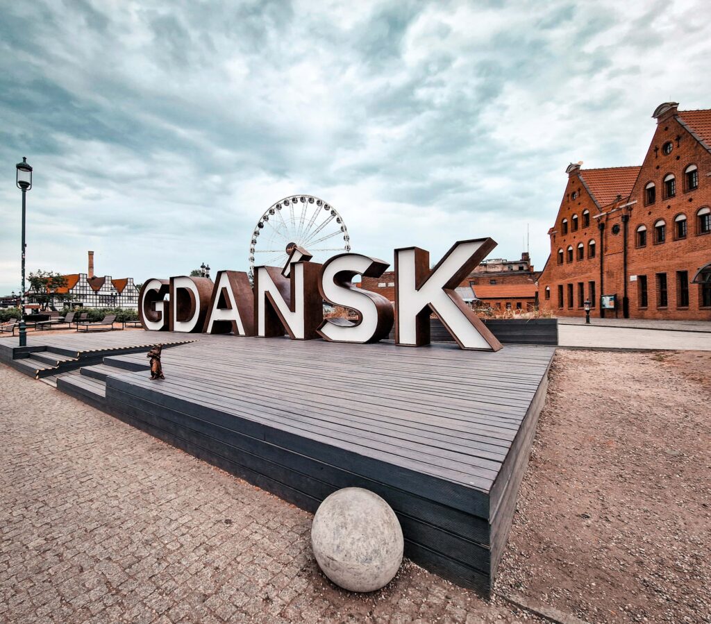 A small platform in the foreground with a "Gdansk" sign sitting on it. There are some buildings in the background to the right and the AmberSky ferris wheel just behind the Gdansk sign. The sky is dark and a little stormy looking.