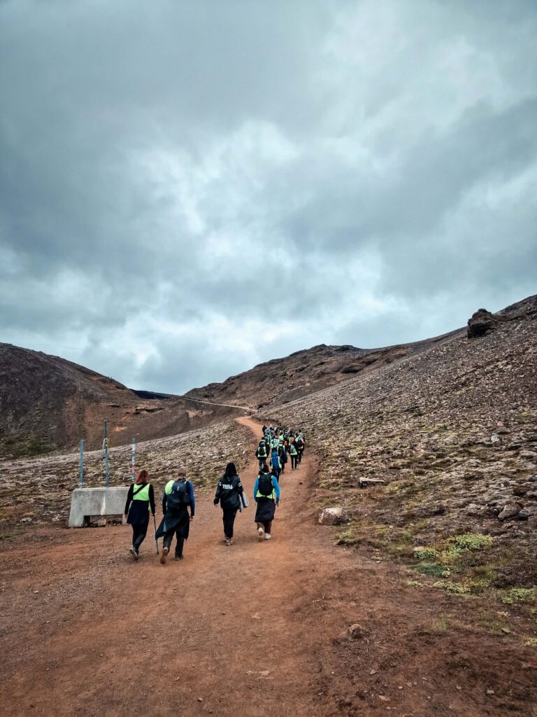 A group of hikers beginning the ascent up a dusty dirt path, with the rocky volcanic landscape on either side of the path. The sky is dark grey and stormy looking.