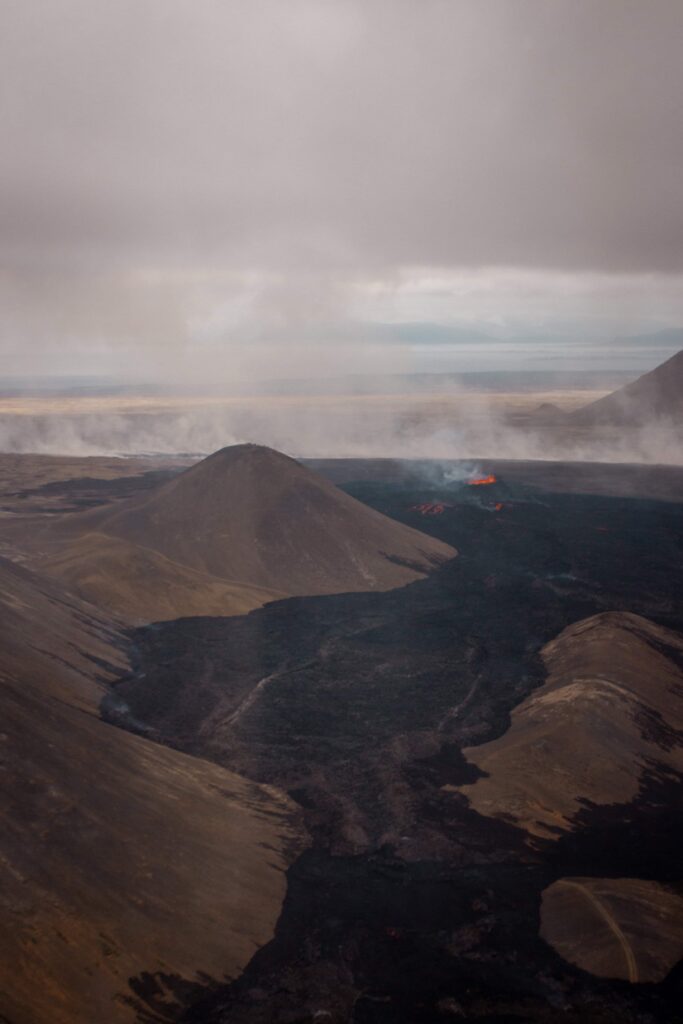 Image from above (via helicopter) showing the lava field of the July 2023 eruption and the Litli-Hrutur crater on the Reykjanes Peninsula (Iceland). The Litli-Hrutur hill is to the left of the new crater, the blackened lava field snakes through the valley in the foreground. Orange lava bubbles from the crater and orange lava flows on parts of the blackened lava field.