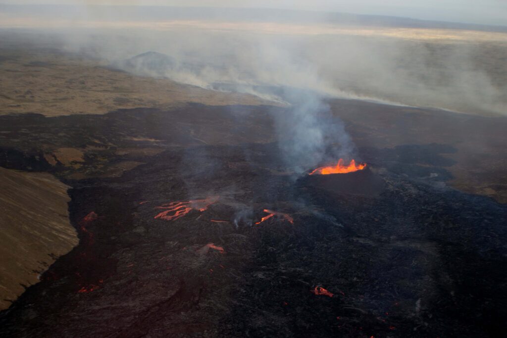 Image from above (via helicopter) showing the lava field of the July 2023 eruption and the Litli-Hrutur crater on the Reykjanes Peninsula (Iceland). The Litli-Hrutur hill is to the left, the blackened lava field is in the foreground and the crater is towards the upper right side. Orange lava bubbles from the crater and orange lava flows on parts of the blackened lava field.